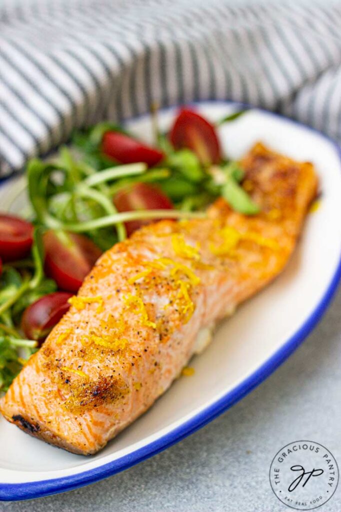 A front view of a salmon fillet on a plate with some greens and tomatoes.