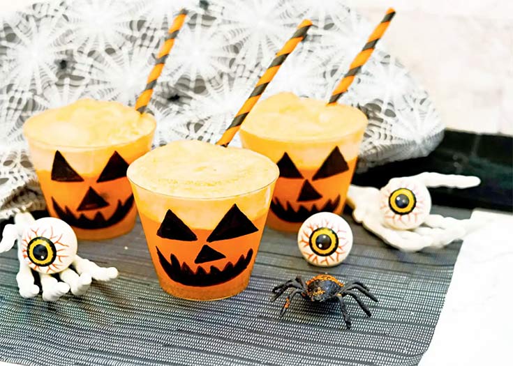Three cups with pumpkin faces and straws sit filled with orange halloween punch.