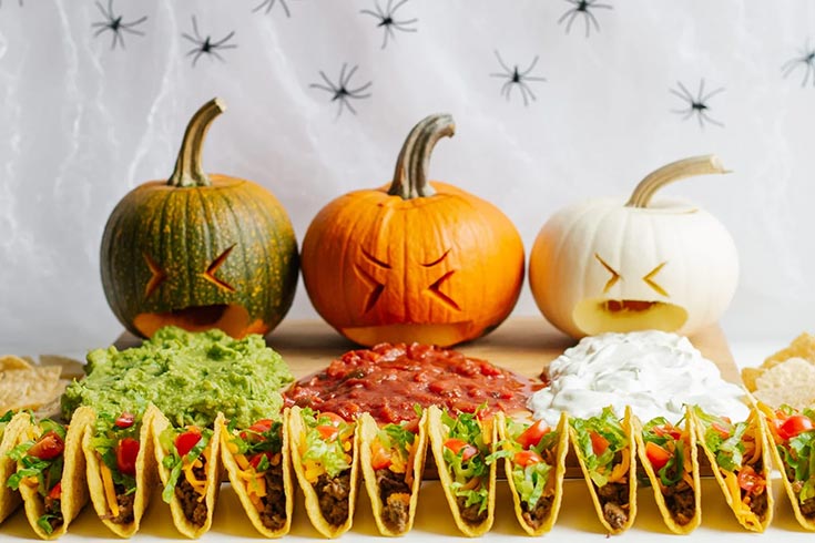 Three pumpkins lined up with faces carved into them to make them look like they are puking. Dip lays in front of them with tacos in front of that.