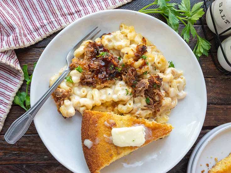 A white plate with a serving of pulled pork mac and cheese. A slice of cornbread lays next to it with a pat of melting butter on top.