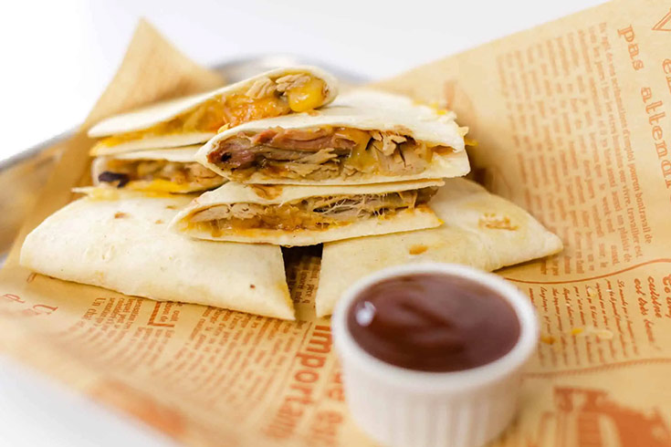 A cut, pulled pork quesadilla lays on parchment next to a small white bowl of dipping sauce.