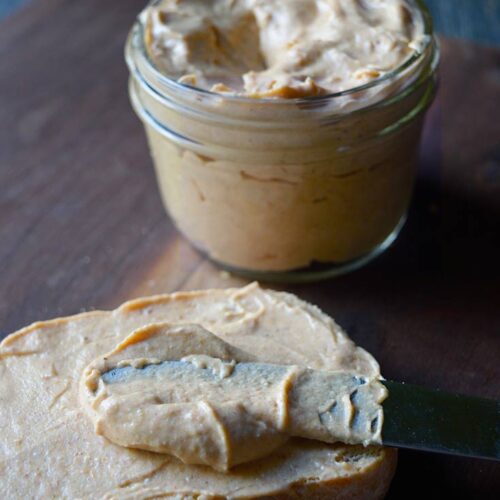 A slice of bread lays on a cutting board covered in pumpkin cream cheese with a jar of the spread sitting behind it.