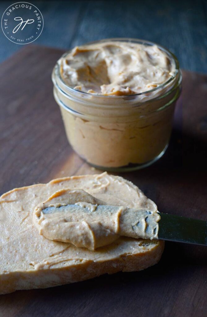 A slice of bread lays on a cutting board covered in pumpkin cream cheese with a jar of the spread sitting behind it.