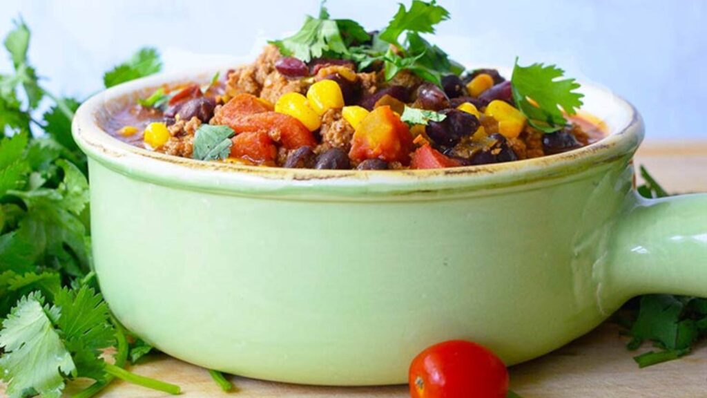 A side view of a light, mint green crock filled with 2 Bean Turkey Chili. The crock sits on a cutting board against a white background.