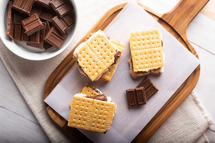 Three s'mores on a parchment-lined cutting board. A bowl of chocolate site next to it.