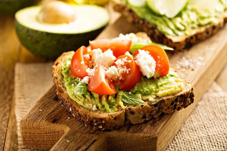 A slice of avocado toast sits on a cutting board. It's garnished with tomatoes and feta cheese.