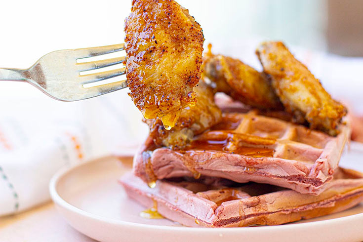 A fork holds some fried chicken from a stack of chicken and red velvet waffles on a plate.