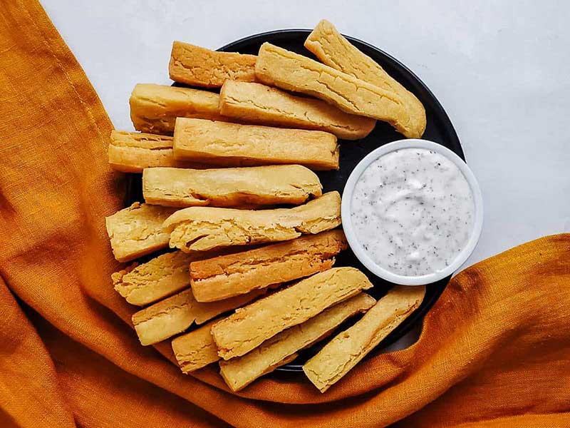 A plate full of chickpea fries with a small bowl of dip all on a black platter.