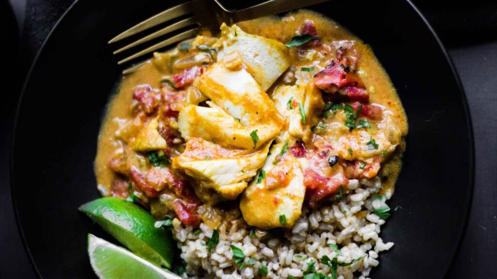 An overhead view of a black bowl filled with coconut tomato curry cod, rice and some lime wedges.