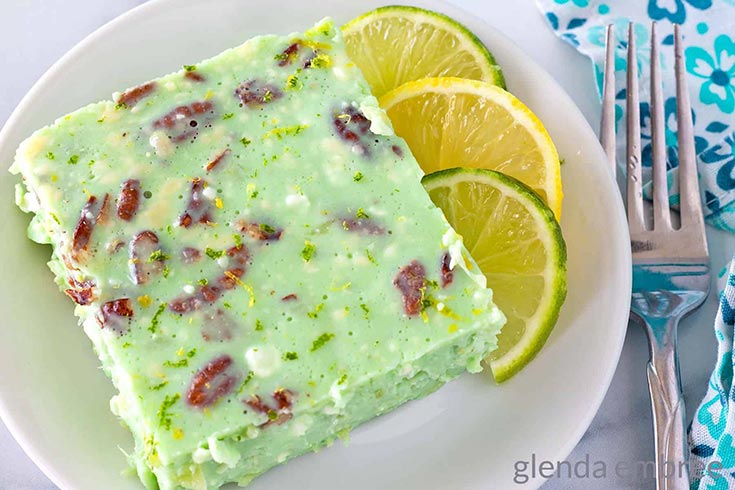 A square slice of lime jello salad on a white plate with some half lime slices.