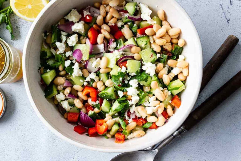 A white mixing bowl holds Mediterranean bean salad. Salad tongs lay next to the bowl.