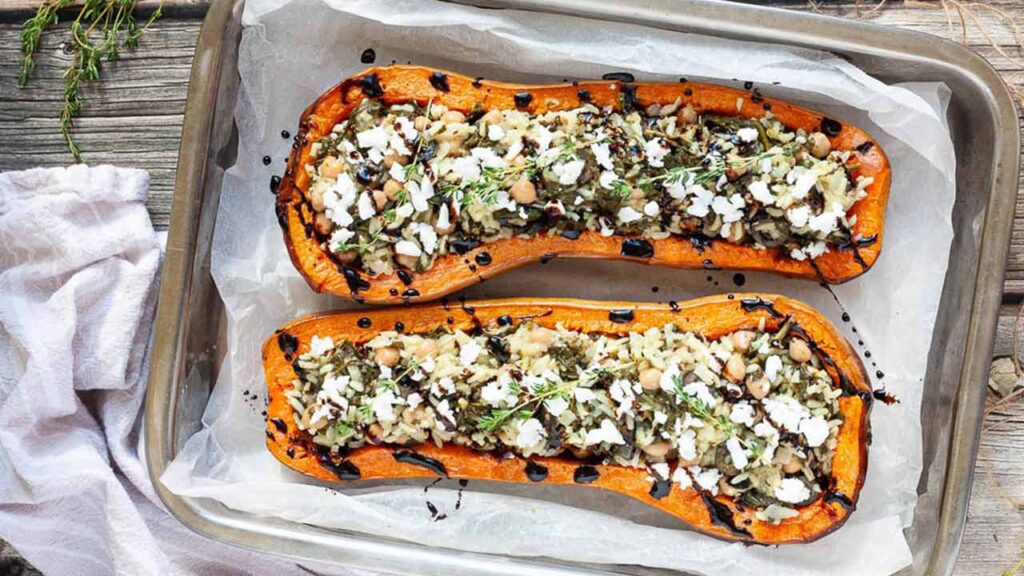 Two stuffed butternut squash halves on a baking sheet. They are garnished with feta crumbles and balsamic glaze.
