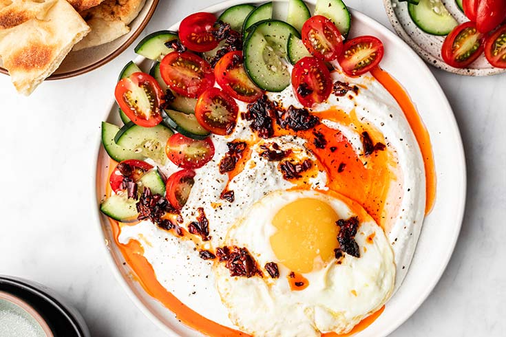 A plate holds a serving of whipped cottage cheese with a fried egg on top and some fresh tomatoes and cucumber slices to the side.