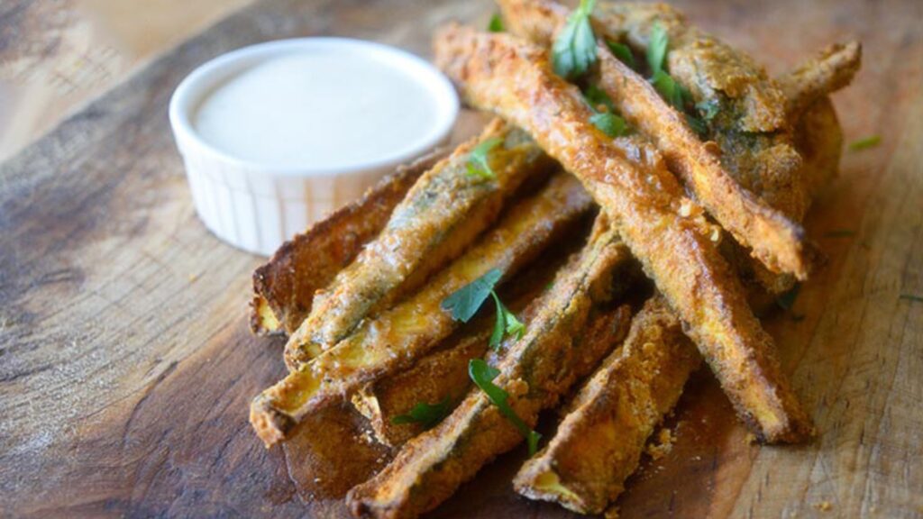 A pile of air fried cajun zucchini sticks laying on a cutting board with a small cup of dip.