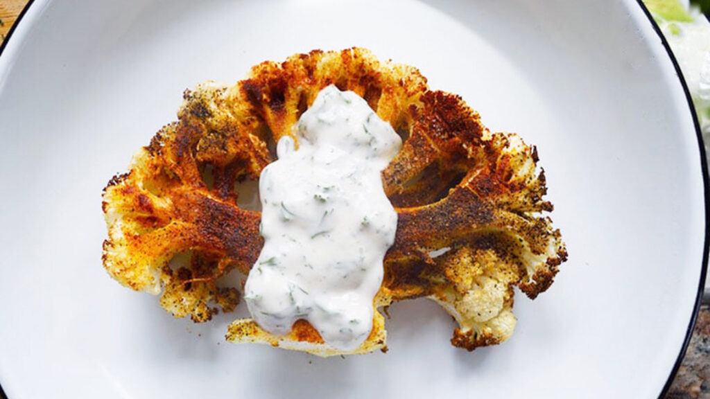 A single cauliflower steak on a plate with yogurt sauce spooned over it.