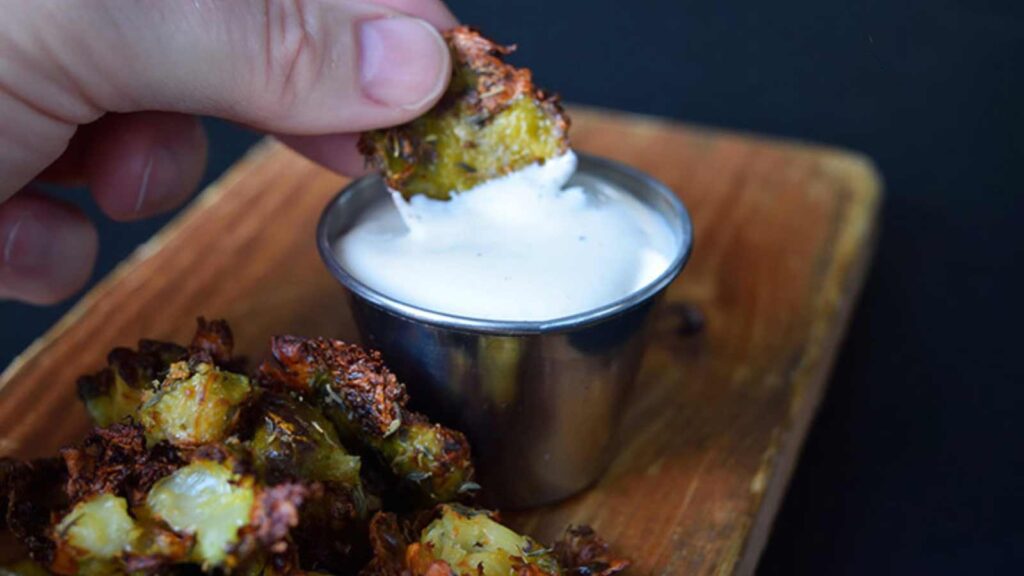 A hand dips a smashed Brussels sprout into a small cup of ranch dressing.