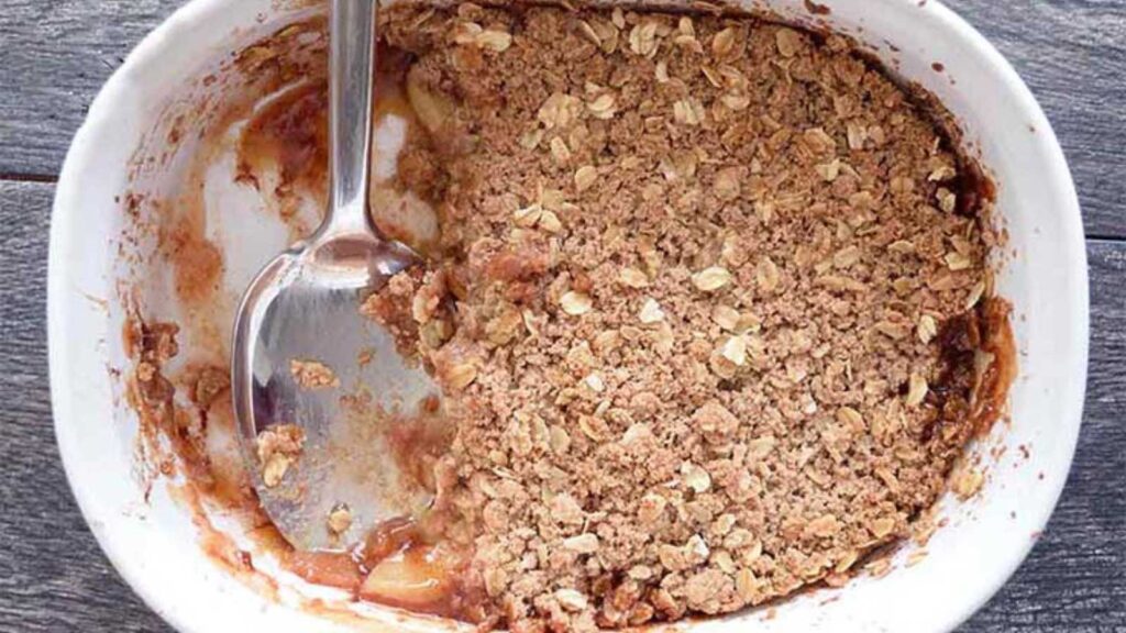 An apple crisp in a white casserole dish with a portion removed. A serving spoon rests in the dish.