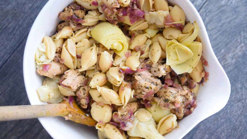 An overhead view looking down into a serving bowl of this delicious artichoke chicken pasta. A wooden spoon is in the bowl, ready to serve the pasta.