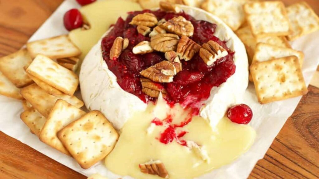 A round of baked brie with cranberry sauce and pecans on top. Crackers surround it.