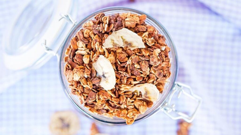 An overhead view of an open jar of banana granola on a purple surface.