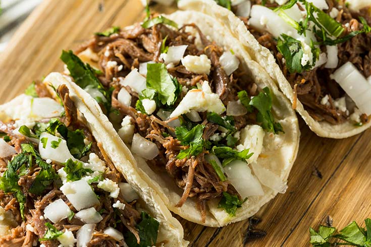 Beef barbacoa tacos in a row on a cutting board.