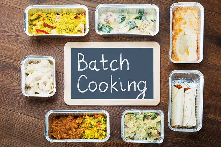 A sign on a table that says, "batch cooking", is surrounded by foil containers of food.
