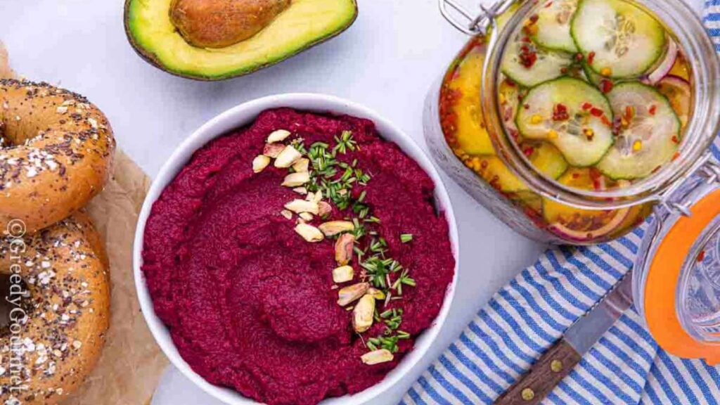 An overhead view of a white bowl filled with beet root hummus. Bagels, a half avocado and a jar of fermented pickles sit around the bowl on a white surface.
