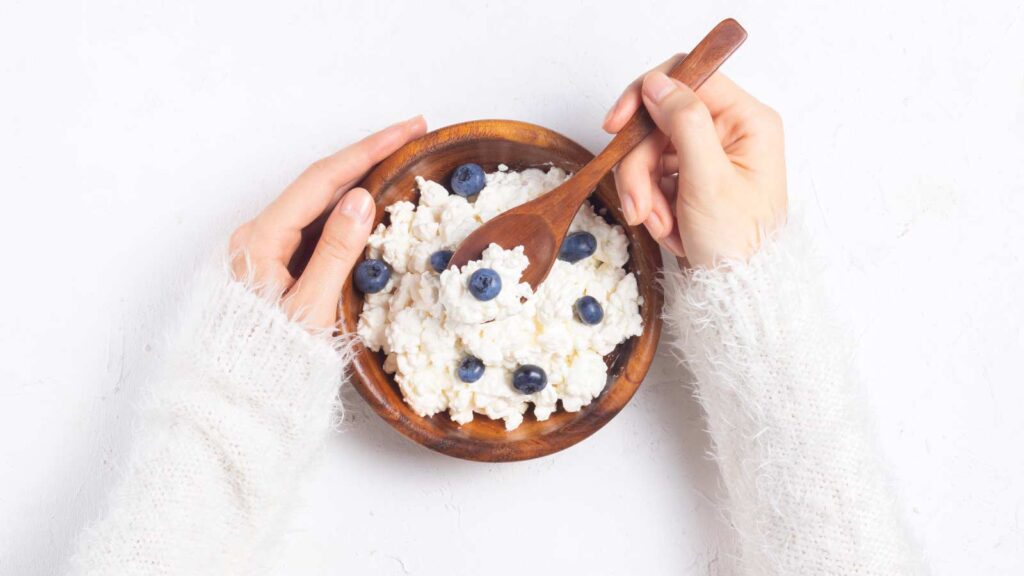 Two hands hold a wooden bowl filled with cottage cheese and blueberries.