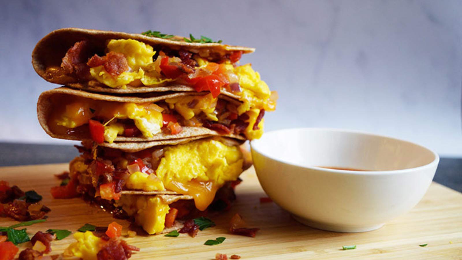 A stack of a cut breakfast quesadilla on a cutting board next to a white bowl of salsa.