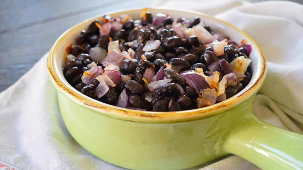 A green crock full of Butter Black Beans sits on a white towel on a countertop.