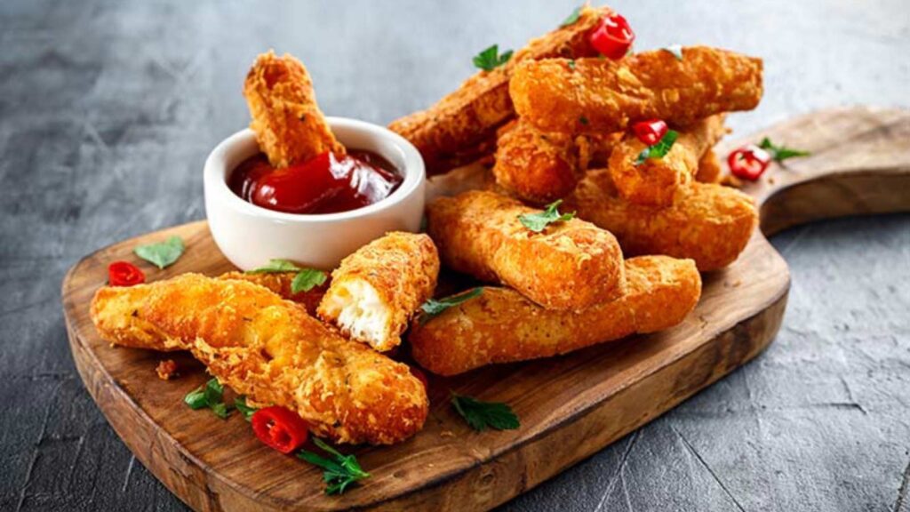 A cutting board holding a pile of cheese sticks and a small white bowl of marinara dip.