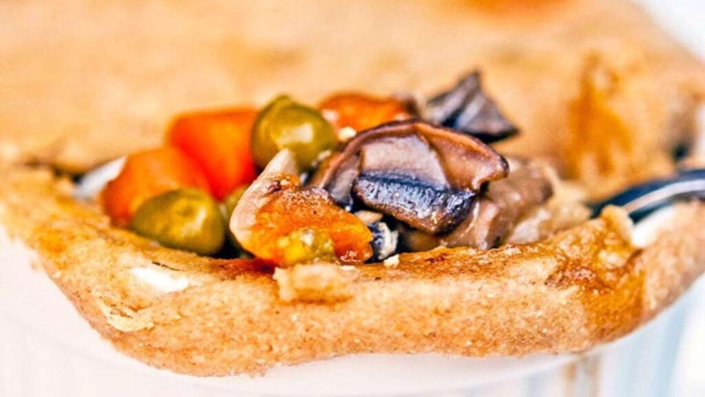 A closeup of a chicken pot pie in a white ramekin. The front of the pie is broken open with a spoon to show mushrooms, peas and carrots on the inside.