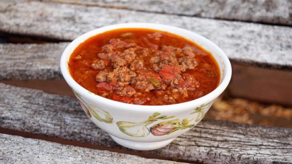 A white, decorative bowl on a wooden surface, filled with Chili Con Carne.