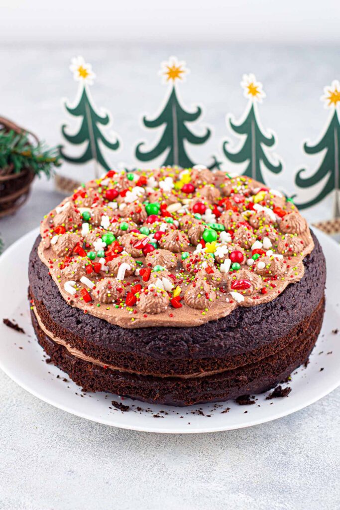 A whole Chocolate Christmas Cake on a platter next to some cutout decorations of Christmas trees.
