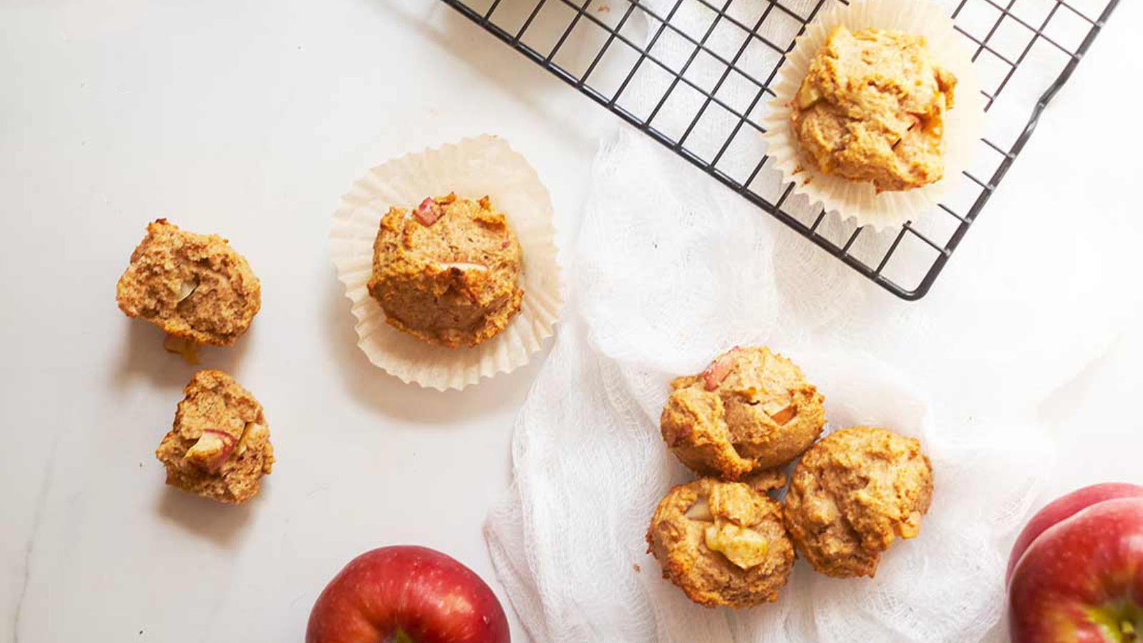 An overhead view looking down on some cinnamon apple muffins and some apples on a white surface.