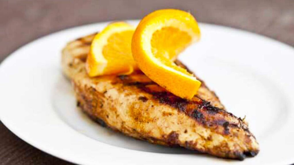 A single Citrus Grilled Chicken breast on a white plate, garnished with an orange twist.