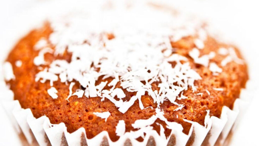A closeup of a muffin in white paper topped with white coconut shreds.