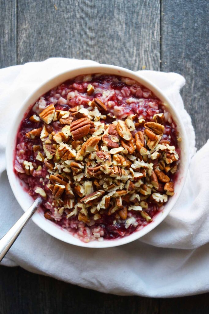 An overhead view of Cranberry Oatmeal in a white bowl with a spoon, topped with pecans.