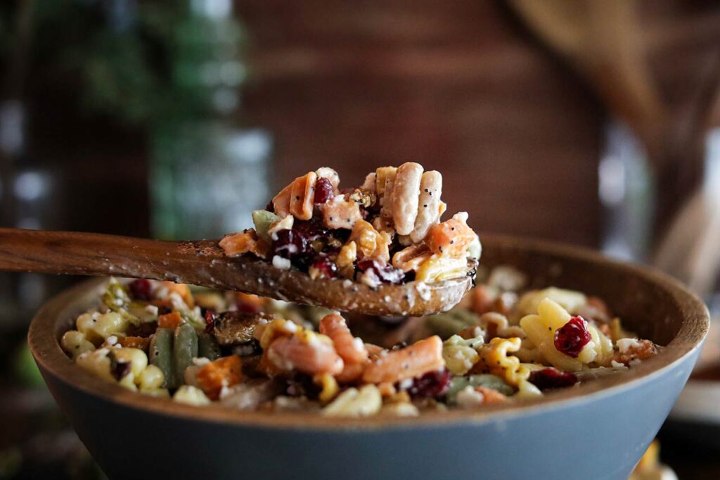 A side view of a wooden spoon lifting up some Fall Harvest Pasta Salad our of a mixing bowl.
