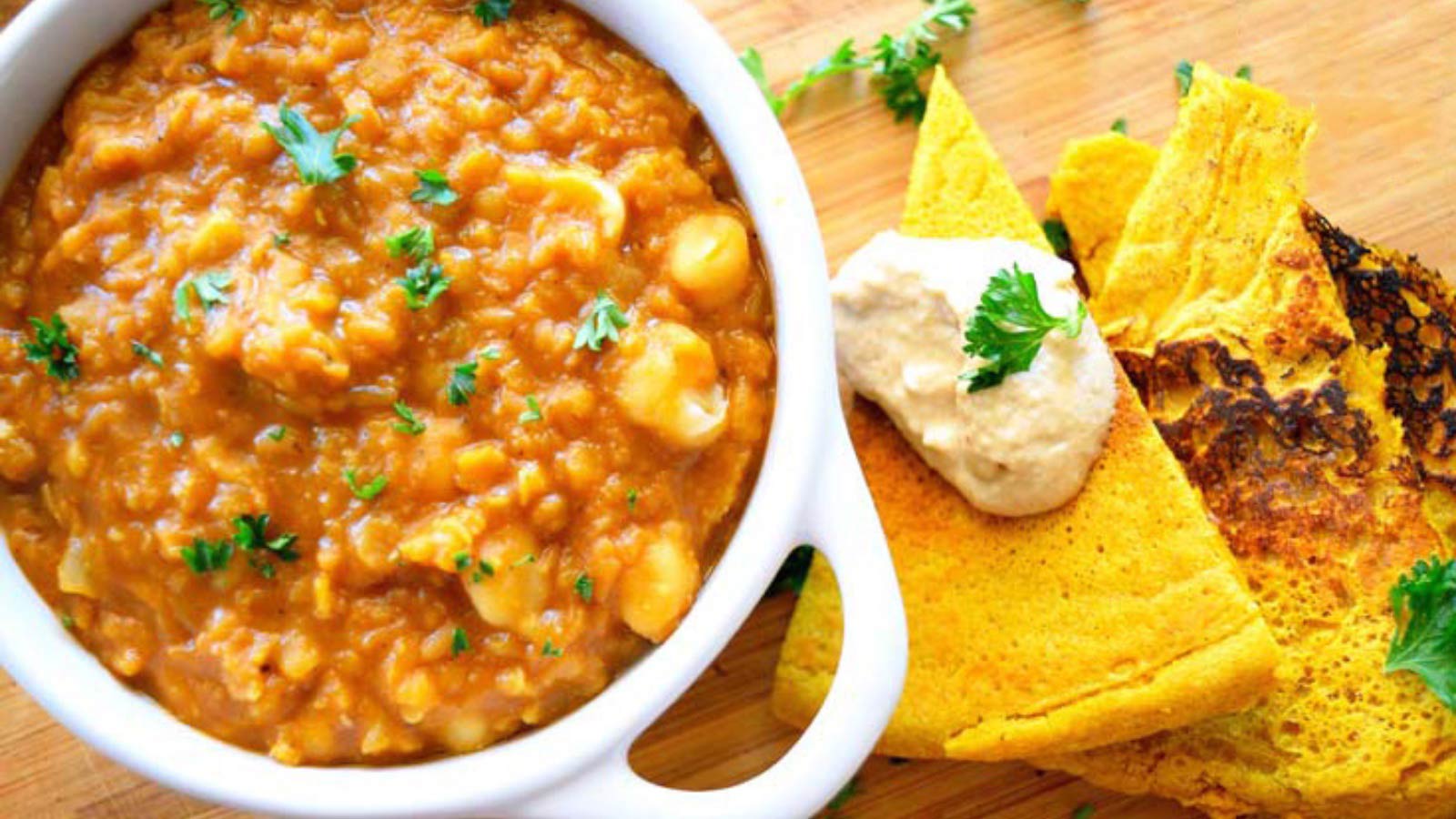 A white crock filled with Garam Masala Red Lentil Stew sits on a cutting board with some flatbread pieces.