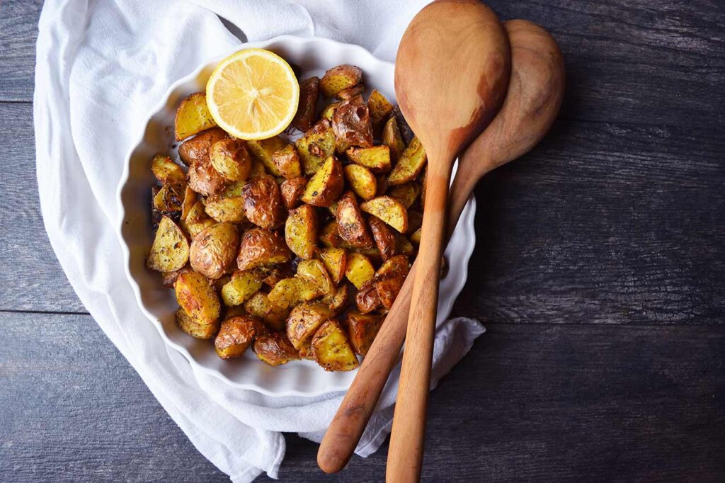 An overhead view of a white dish filled with Garlic Lemon Potatoes and half a lemon. Wooden spoon rest on the dish.