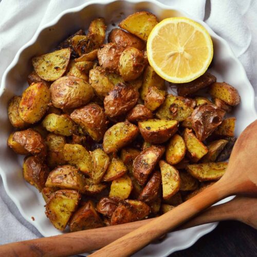 An overhead view of a white dish filled with Garlic Lemon Potatoes. A half lemon is wedged into the side of the dish.