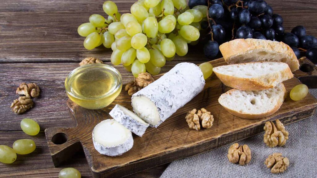 A cutting board with a log of goat cheese, some bread slices and some grapes.