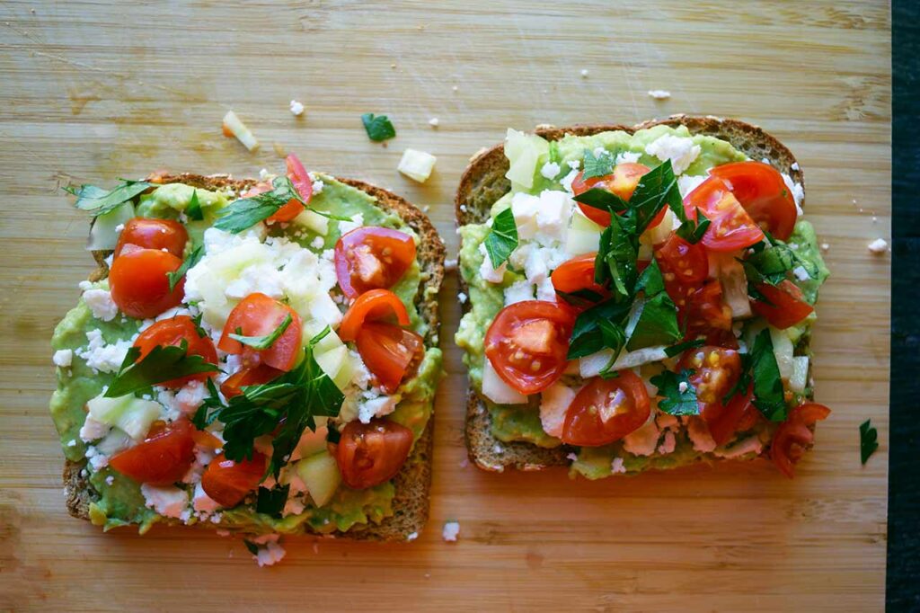 Two slices of Greek avocado toast laying side by side on a cutting board.