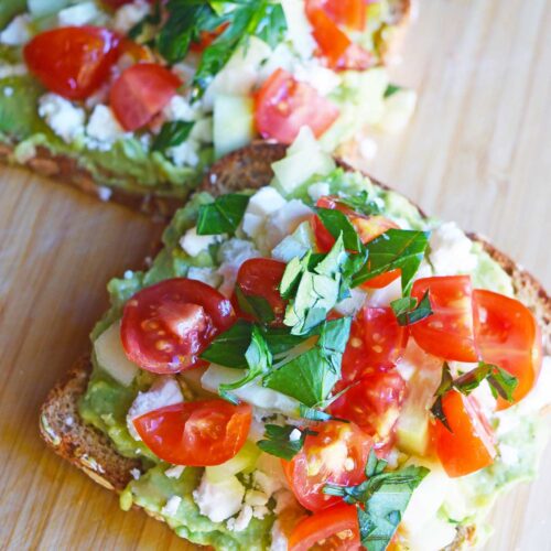 A closeup of two slices of Greek avocado toast on a cutting board.