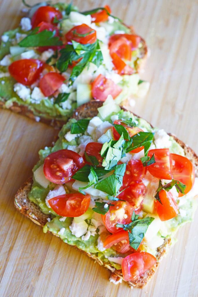 A closeup of two slices of Greek avocado toast on a cutting board.