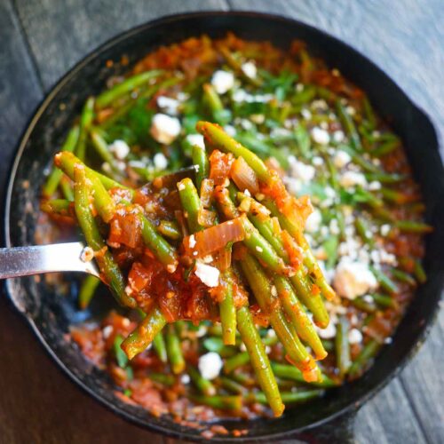 A serving spoon lifts some Greek green beans out of a cast iron skillet.