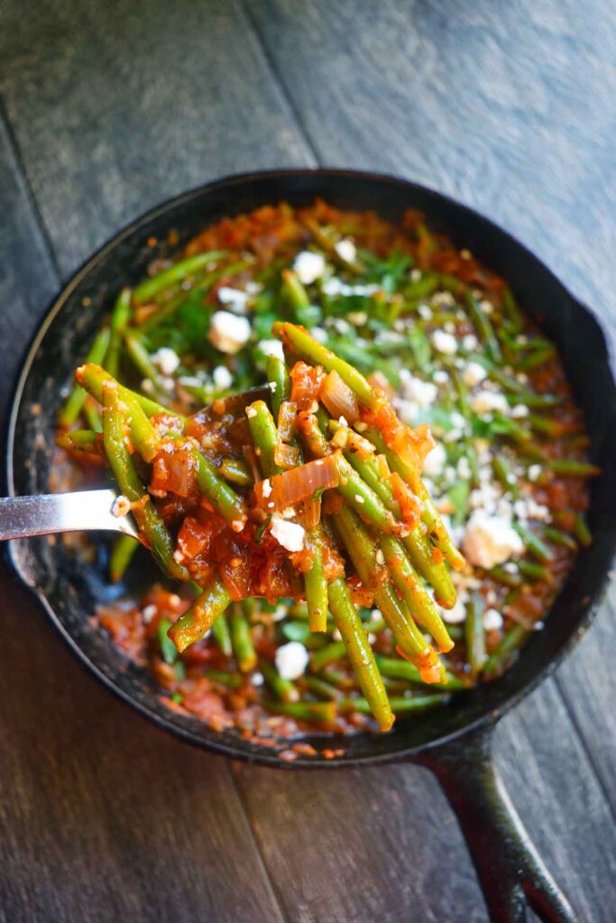 A serving spoon lifts some Greek green beans out of a cast iron skillet. A great side dish for a Mediterranean Diet Thanksgiving Menu.