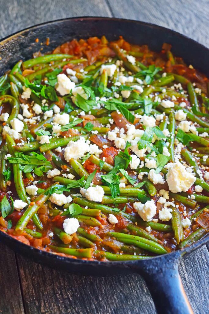 Greek green beans in a cast iron skillet garnished with feta cheese crumbles and fresh, chopped parsley.