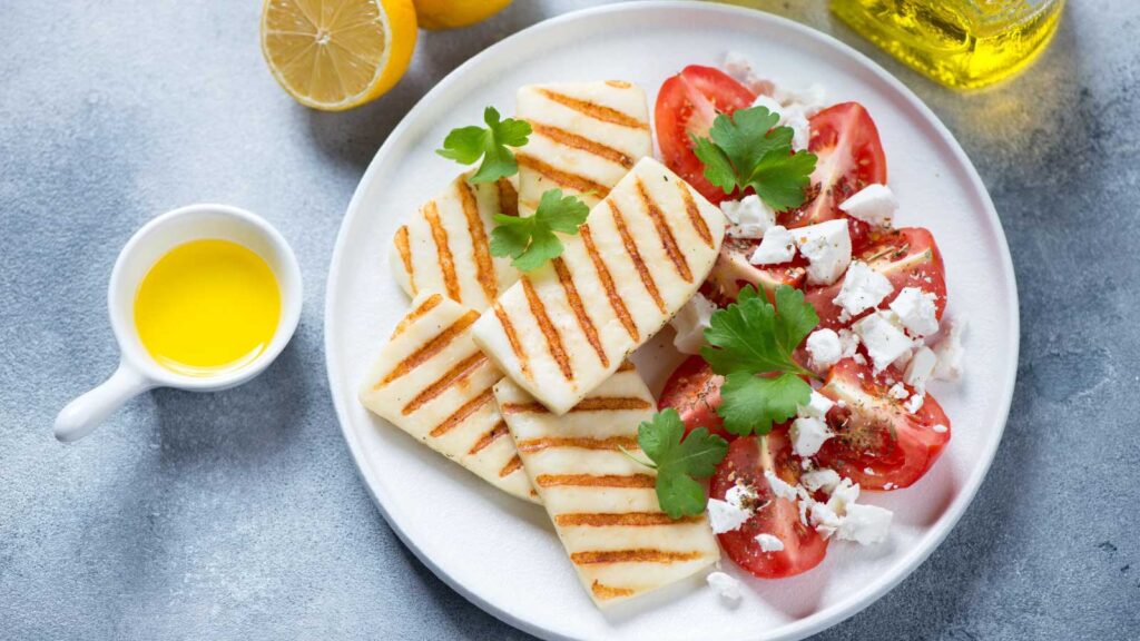 A white plate holds Halloumi cheese slices that have been grilled and some fresh tomatoes with feta crumbles sprinkled over them.